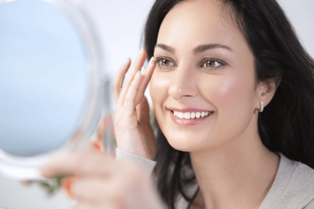 Smiling woman looks at herself in a handheld mirror while touching the outer corner of her eye