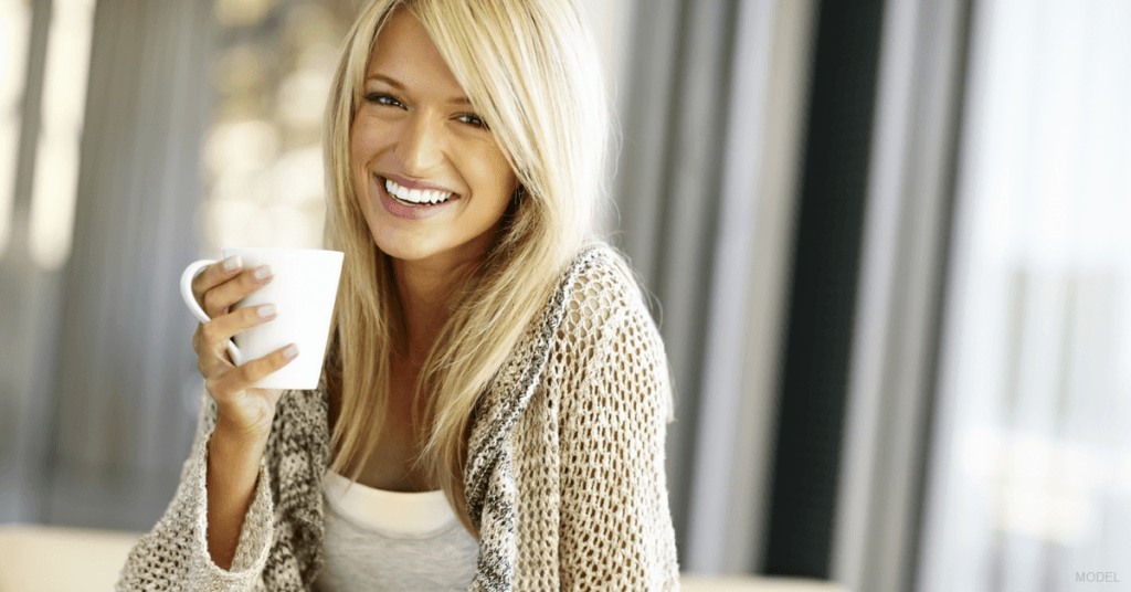 A woman sits on a bed, sips coffee and considers her mommy makeover options.