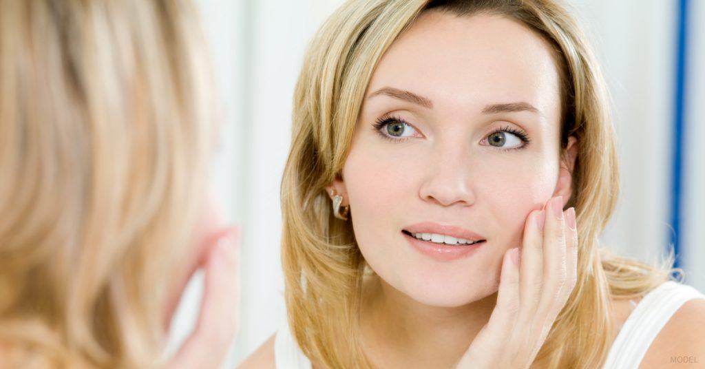 A woman looks in the mirror and touches her face as she is beginning to see the positive results of RF Microneedling.