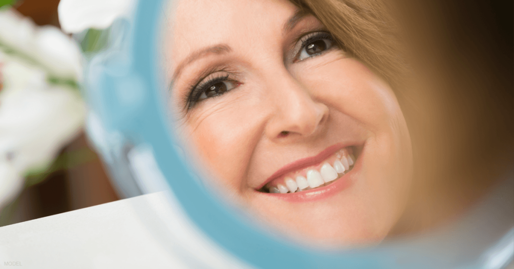 A woman looks in the mirror with delight after recovering from her eyelid surgery.