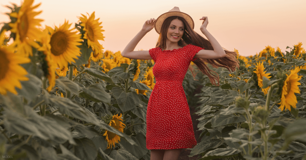 A woman in a red dress walks through a field of sunflowers after receiving nonsurgical mommy makeover treatments.