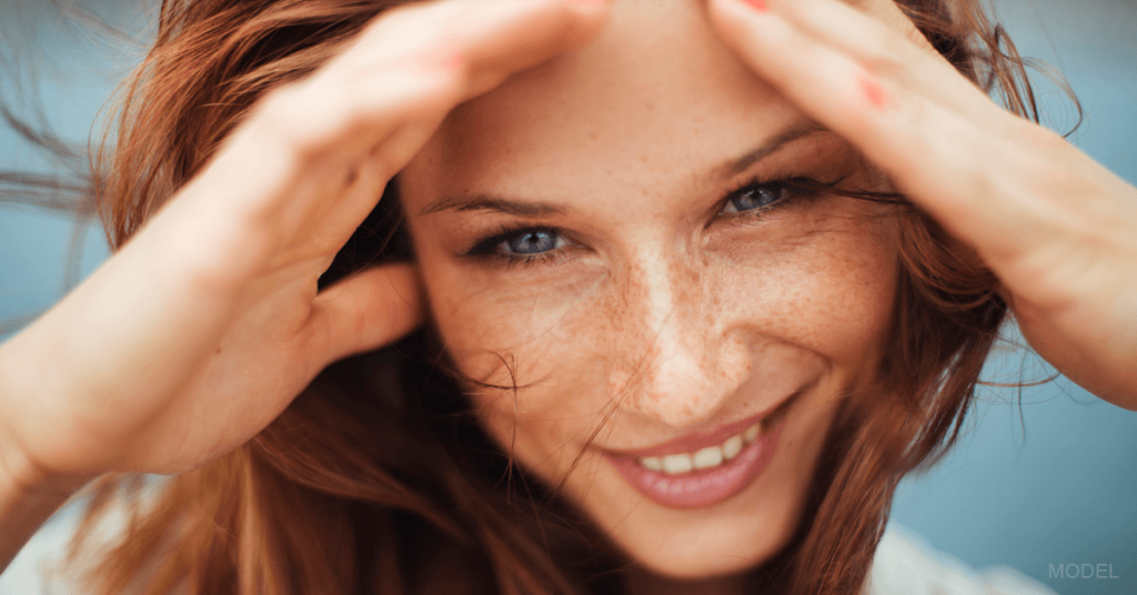 A woman who has received BOTOX injections smiles and shades her face from the sun with her hands.