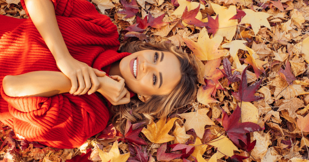 A smiling woman in a red sweater rests on her back among fallen autumn leaves.