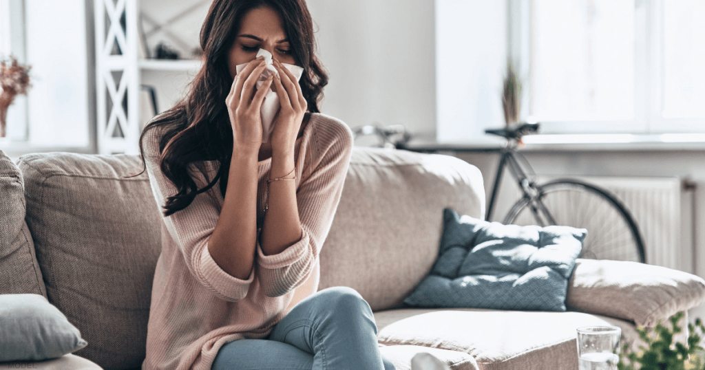 A woman sits on a sofa and blows her nose into a tissue.