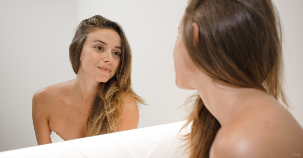 Woman looking in the mirror to determine if plastic surgery or nonsurgical procedures are best for her (model)